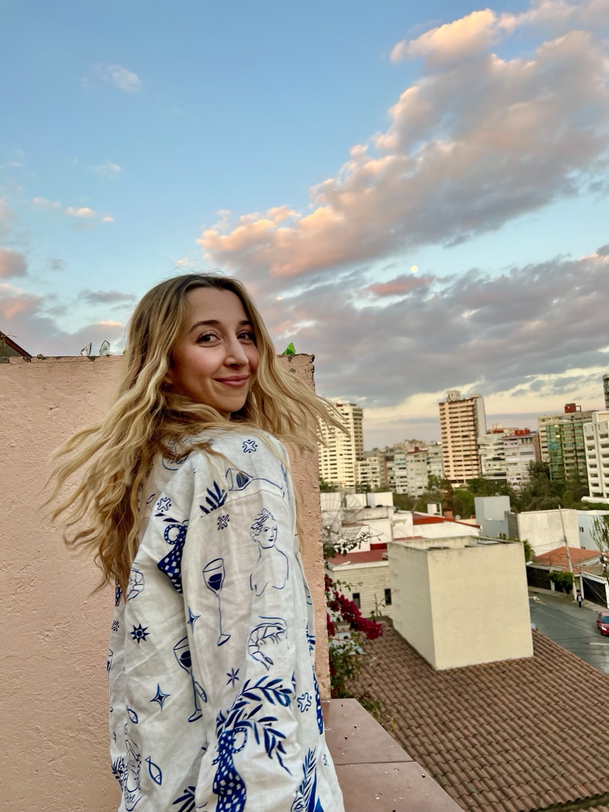 Aislinn on a rooftop at sunset, smiling over her shoulder, pastel clouds and a city behind her